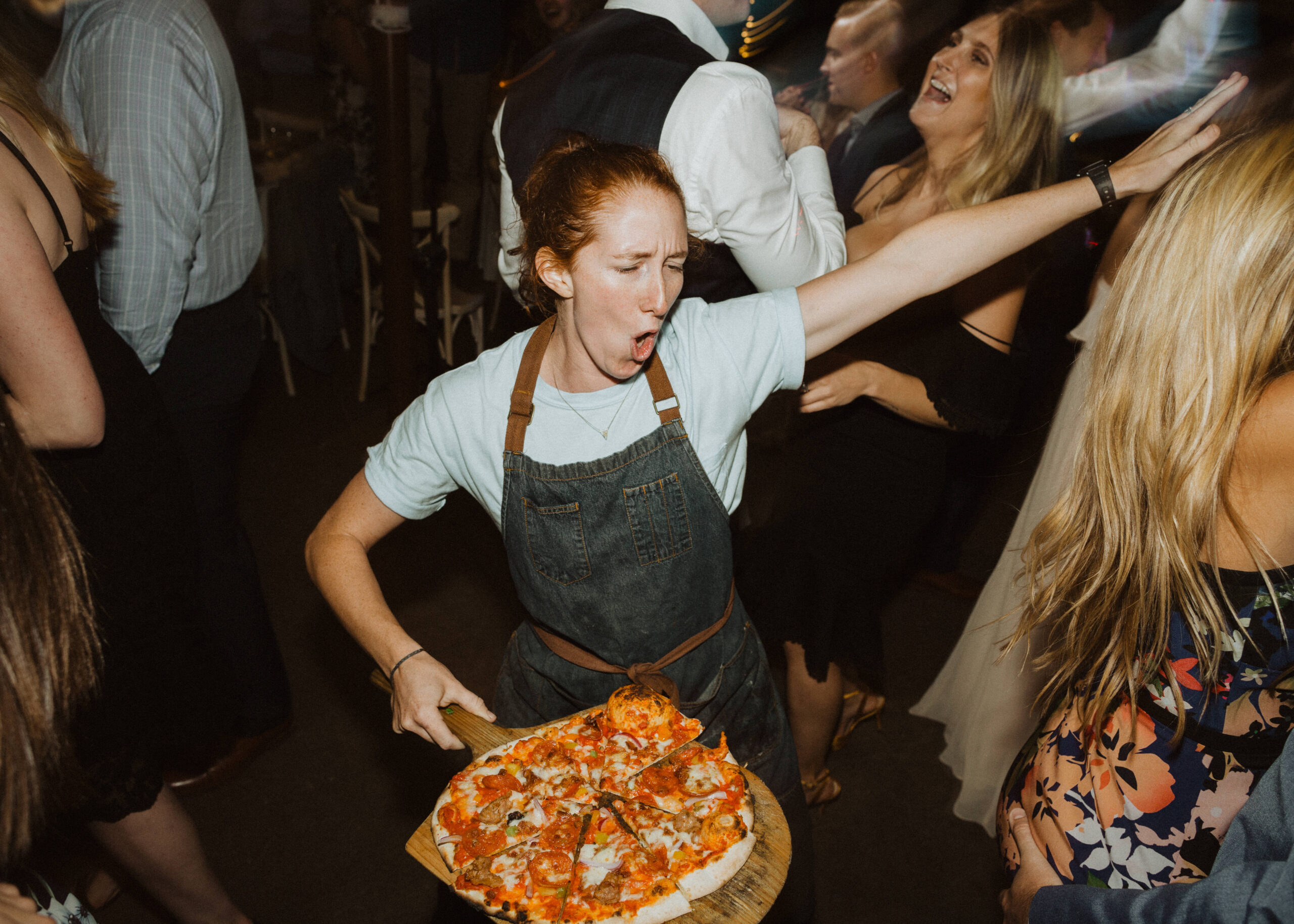 A woman in an apron holding a pizza at a party.
