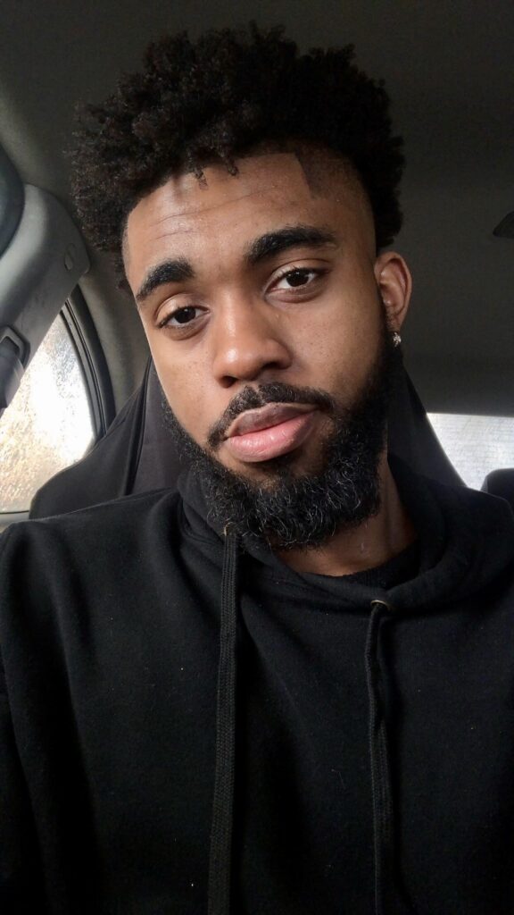 A black man sitting in a car with a beard, on his way to provide DJ service at a Georgia wedding.