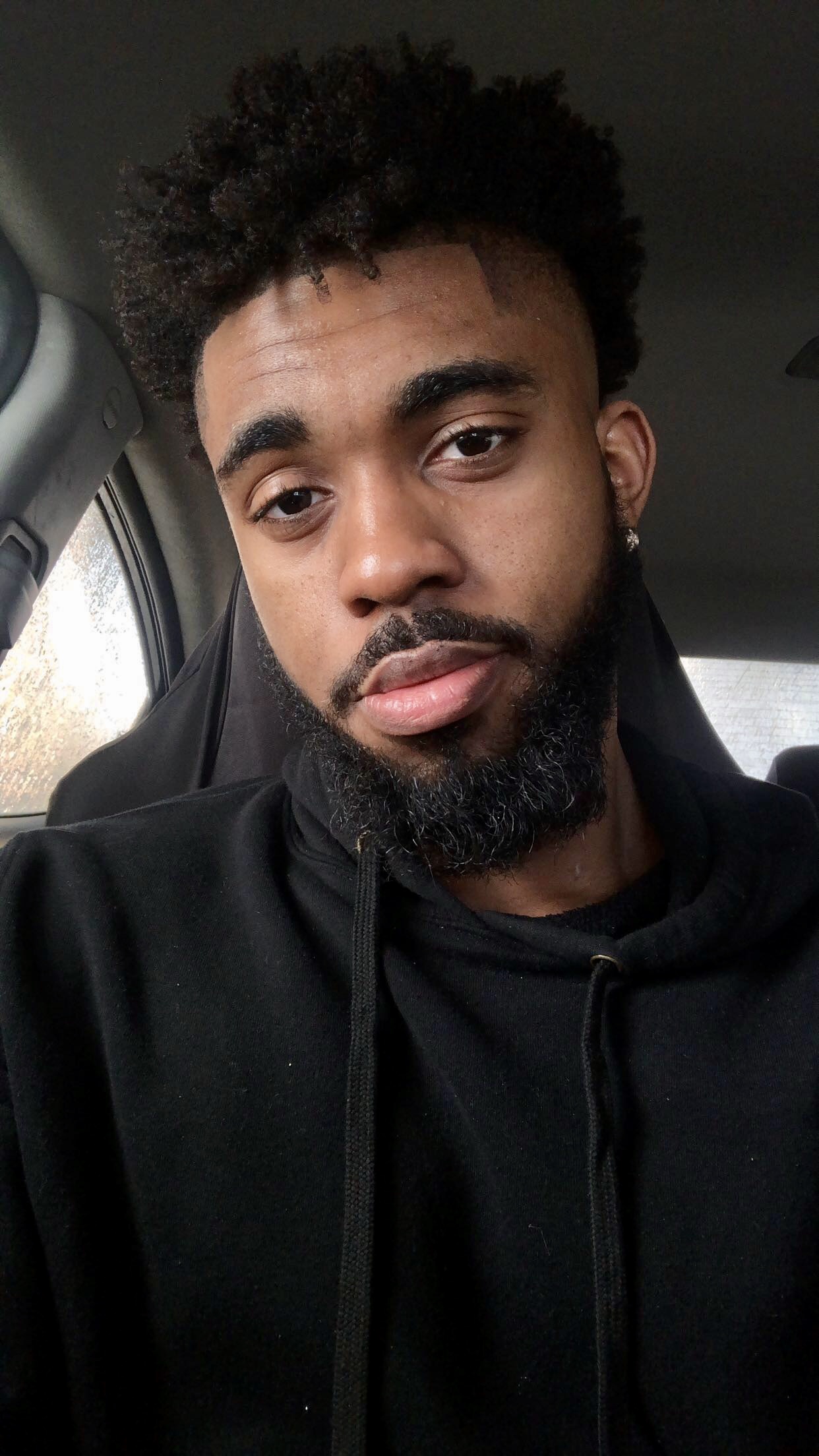 A black man sitting in a car with a beard, on his way to provide DJ service at a Georgia wedding.