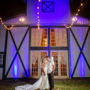 A bride and groom standing in front of a barn at night, with a Georgia wedding DJ setting up in the background.