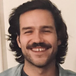 A smiling person with long hair and a mustache, wearing a light-colored shirt, stands in front of a serene backdrop. The cheerful expression on their face radiates warmth and positivity.