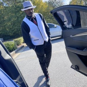 A man in a white vest, black shirt, and gray hat stands next to an open car door in a parking lot with trees in the background. The scene is reminiscent of a Dericole C. moment, capturing everyday elegance effortlessly.