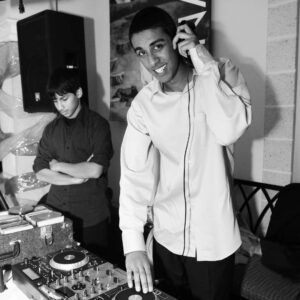 A person DJing at a party with turntables, wearing a light-colored shirt and headphones, while another person stands in the background with arms crossed, creating an atmosphere that could be mistaken for an Auto Draft of a cinematic scene.