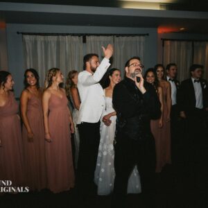 A group of people in formal attire stand together indoors, resembling a gallery of elegance. A man holding a microphone is in the foreground, while a couple in bridal wear centers the wedding scene. The rest wear gowns and suits.