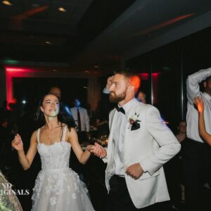 A bride in a white dress and a groom in a white jacket dance at their wedding reception, surrounded by guests, creating a beautiful gallery of memories.