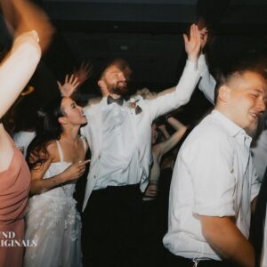 A lively and joyful wedding reception is in full swing as a group of people, including the bride and groom, dance with raised arms in their formal attire. The scene unfolds like a vibrant gallery of celebration.