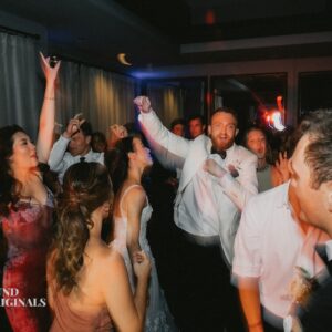 A group of people in formal attire enthusiastically dancing at a lively wedding, with colorful lights creating a festive atmosphere.