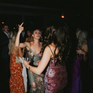 A group of people in formal attire dance and socialize at an indoor event, reminiscent of a wedding reception. Some hold drinks, while one woman raises her arm. The dimly lit setting creates a charming ambience with the "Sound Originals" logo subtly displayed in the bottom corner.