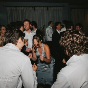 A group of people in formal attire dance and socialize at an indoor wedding event with dim lighting.
