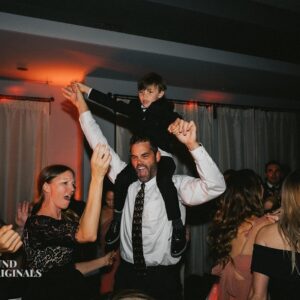 An adult man with a child on his shoulders, both raising their hands, amid a group of joyful people dancing in a dimly lit wedding reception.