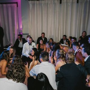 A large group of people seated and socializing in a dimly lit wedding gallery with white curtains and purple lighting.