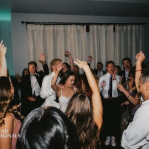 A bride and groom dance surrounded by guests at a wedding reception, with several people raising their hands and smiling. The atmosphere appears lively and celebratory, resembling a joyful gallery of love and happiness.