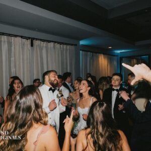 A group of formally dressed people are gathered, laughing and singing, in a dimly lit room with white curtains. Some hold drinks, and one person is pointing. The atmosphere appears festive and lively, much like a wedding reception in an intimate gallery setting.