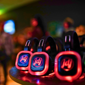 Three pairs of glowing headphones sit on a table in a dimly lit room, ready for the ultimate Silent Disco experience, each displaying the logo "jdj" on their ear cups.