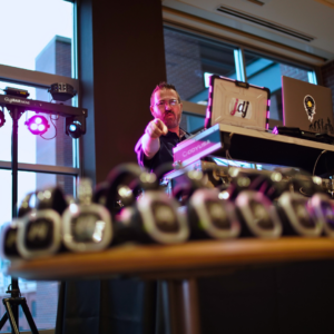 The DJ at a setup table points towards the camera with headphones in the foreground, capturing the vibrant atmosphere of a Silent Disco under colorful lighting.