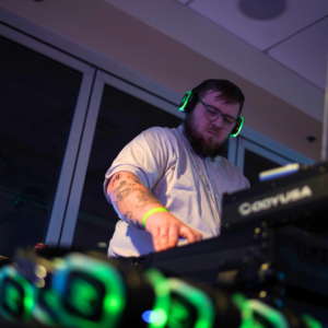 A person wearing headphones is DJing at a mixing console with green illuminated accents, creating an electrifying ambiance for the silent disco in a dimly lit room.