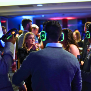 At a vibrant party adorned with colorful lights, guests are immersed in the unique experience of a silent disco. Each person, sporting headphones and holding drinks, dances to their own rhythm, creating a lively atmosphere without the usual blare of speakers.