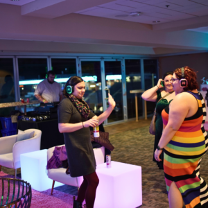 At a vibrant silent disco, people donning light-up headphones mingle and dance in a stylish lounge, with modern decor framing the backdrop and a DJ spinning tracks in the background.