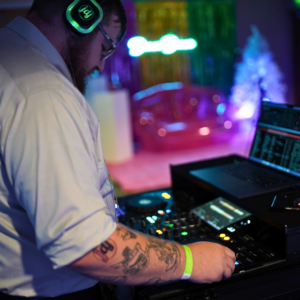 A DJ sporting a white shirt and tattoos on his arm expertly navigates the mixing console, setting the tone for a vibrant silent disco. Neon lights pulse in the background, casting an electrifying glow over his headphones and laptop.