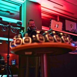 A DJ stands behind turntables and equipment on a table, orchestrating the music under vibrant lighting. In the foreground, a tray of small cups is perfectly placed to elevate the event's ambiance.