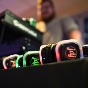 A row of brightly lit headphones on a table hints at an upcoming Silent Disco, with a blurred person and equipment in the background adding to the anticipation.