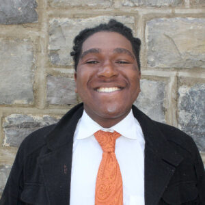 A person in a suit and orange tie smiles in front of a stone wall.
