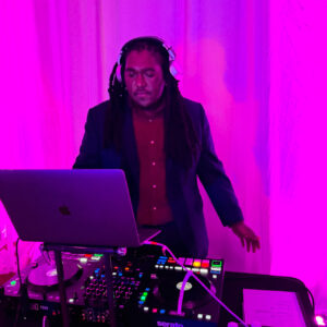 A person with headphones DJs using a laptop and controller on a table with a vibrant pink and purple backdrop.