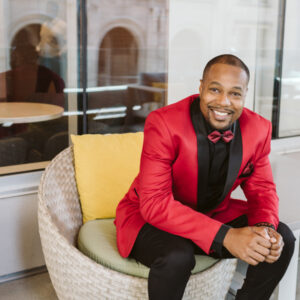 Man in a red jacket and bow tie, sitting and smiling, with glass windows in the background.