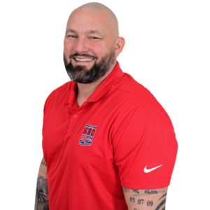 A man with a beard and tattoos, wearing a red Nike polo shirt with a logo on the chest, stands against a plain white background, smiling at the camera—perfectly capturing the approachable vibe of professional DJs for event entertainment.