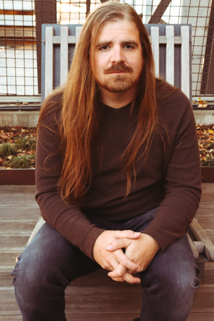 A man with long brown hair and a beard sits on a wooden chair outdoors, wearing a dark sweater and jeans, with hands clasped in his lap.