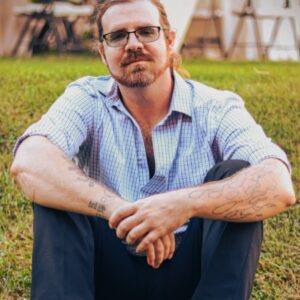 A man with glasses and tattoos sits on grass with his arms resting on his knees, wearing a light plaid shirt and dark pants. In the blurred background, tables and chairs hint at an event music setting provided by Professional DJs.