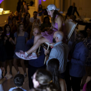 A young woman in a pink dress and cap is lifted on a chair by several people at a crowded indoor party, as guests cheer and watch—capturing the joyful spirit often seen in Bat Mitzvah pictures.