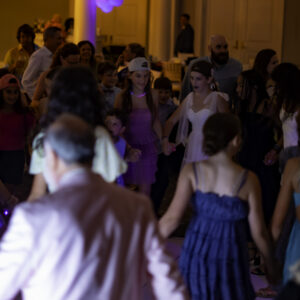 A large group of people, mostly children, stand in a circle holding hands at an indoor event with dim lighting, creating a memorable moment perfect for Bar Mitzvah pictures.