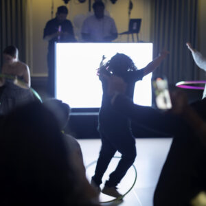 People dance with hula hoops in front of a DJ booth at a dimly lit event, with a brightly lit screen in the background—capturing the fun energy seen in memorable Bar Mitzvah pictures.