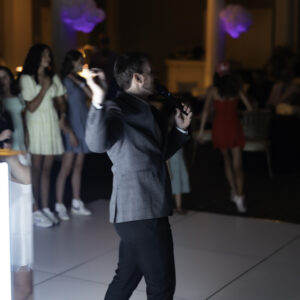 A man in a gray blazer speaks into a microphone on a lit dance floor, facing a group of people, capturing the lively atmosphere perfect for memorable Bar Mitzvah pictures, with tables and chairs in the background.