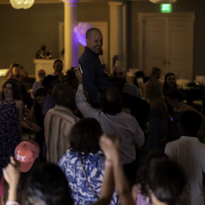 A group of people gather and dance at an indoor event while several individuals lift a smiling man above the crowd, capturing the joyful energy often found in memorable Bar Mitzvah pictures.