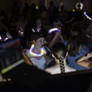 A group of young people wearing glow necklaces gather and socialize in a dimly lit room, with colorful lights and a blurred DJ booth in the foreground—capturing the lively spirit often seen in Bat Mitzvah pictures.