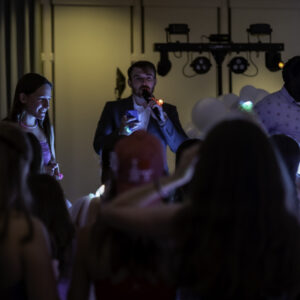A man speaks into a microphone on stage, with another at DJ equipment nearby, as the crowd looks on in a dimly lit room—capturing the energy often seen in vibrant Bar Mitzvah pictures.