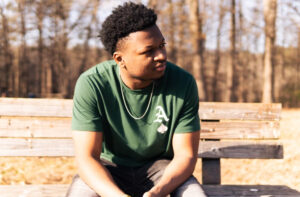 A man in a green t-shirt with a white letter "A" sits on a wooden bench outdoors, looking to his left. Leafless trees and sunlight are visible in the background.