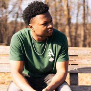 A man in a green t-shirt with a white letter "A" sits on a wooden bench outdoors, looking to his left. Leafless trees and sunlight are visible in the background.