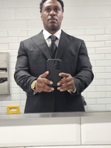 A man wearing a black suit and tie, resembling the polished style of professional DJs, takes a mirror selfie in a tiled restroom while holding a smartphone.