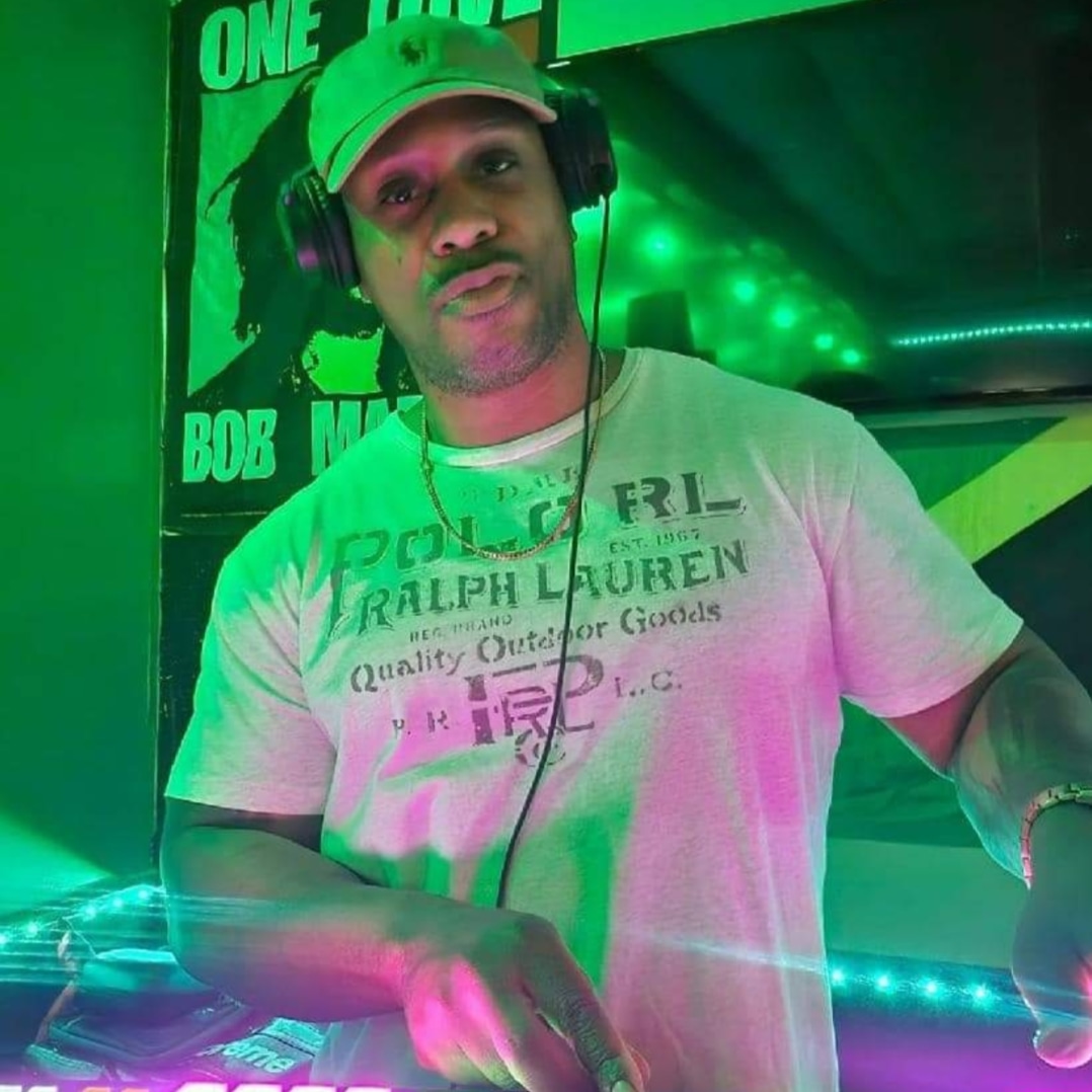 A man wearing headphones and a light-colored Ralph Lauren t-shirt stands at DJ equipment with green and pink lighting; a Bob Marley poster hangs in the background, creating an atmosphere favored by professional DJs.