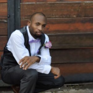 A man in formal attire with a pink rose boutonniere crouches in front of a wooden wall, looking at the camera.