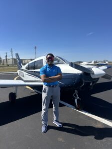 A man wearing sunglasses and a blue shirt stands with arms crossed in front of a small single-engine airplane on an airport tarmac.