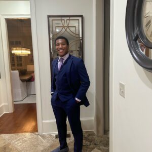 A person in a dark blue suit, light lavender shirt, and matching shoes stands smiling in a hallway with tiled flooring and framed wall décor.
