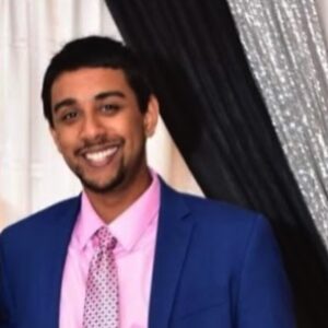 A man in a blue suit, pink shirt, and patterned tie smiles while standing in front of black and white draped curtains, ready to entertain as one of the top professional DJs.