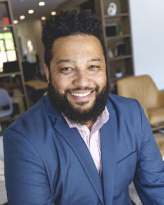 A man with a beard and curly hair, wearing a blue suit and pink shirt, smiles with energy and creativity while seated in a modern office space.