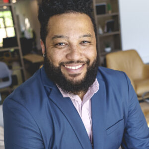 A man with a beard and curly hair, wearing a blue suit and pink shirt, smiles with energy and creativity while seated in a modern office space.