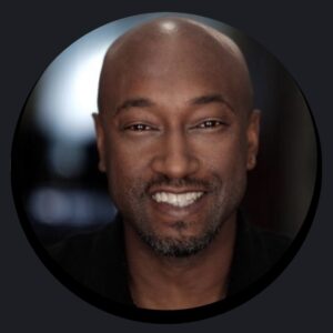 A bald man with a close-cropped beard and mustache, resembling many professional DJs, smiles at the camera against a softly blurred background.