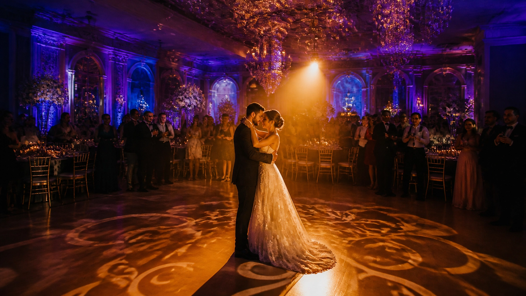 A couple shares their first dance in the center of a decorated ballroom with guests watching and colorful lighting illuminating the scene.
